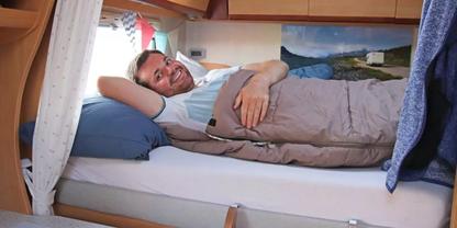 Ein l&auml;chelnder Mann liegt auf der Seite in einem Bett in einem Wohnmobil, mit Kopfkissen und Decke; Holzm&ouml;bel, ein Fenster mit Vorhang und ein Landschaftsbild an der Wand sind sichtbar
