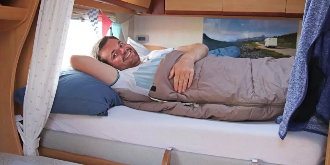 Ein l&auml;chelnder Mann liegt auf der Seite in einem Bett in einem Wohnmobil, mit Kopfkissen und Decke; Holzm&ouml;bel, ein Fenster mit Vorhang und ein Landschaftsbild an der Wand sind sichtbar