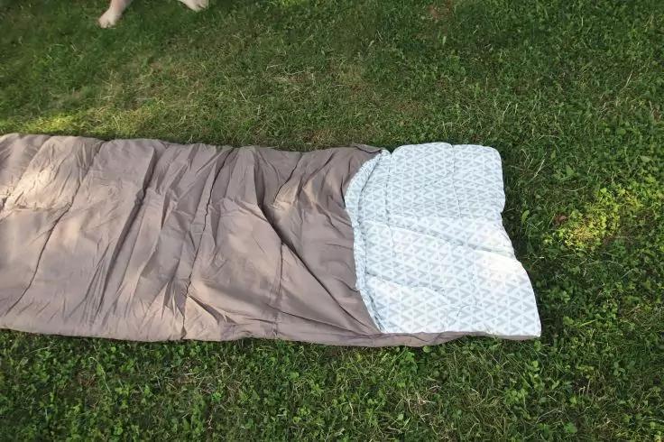 Brauner Schlafsack mit aufgeschlagener wei&szlig; gemusterter Decke auf gr&uuml;nem Gras; oben links teilweise wei&szlig;e Pfoten sichtbar