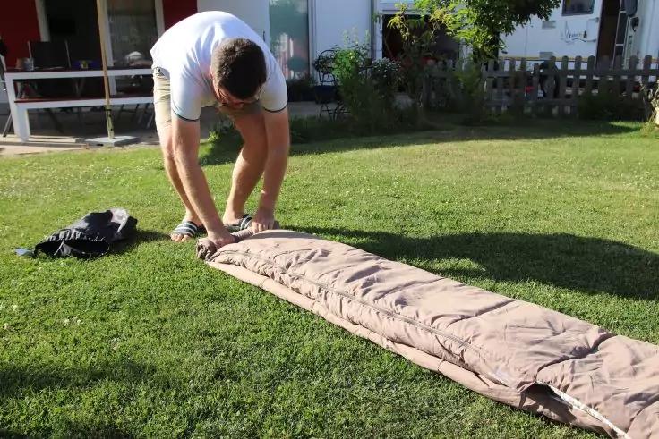 Person rollt auf gr&uuml;nem Rasen einen langen, hellen Schlafsack aus; Jacke liegt daneben; Haus mit Terrasse und Pflanzen im Hintergrund