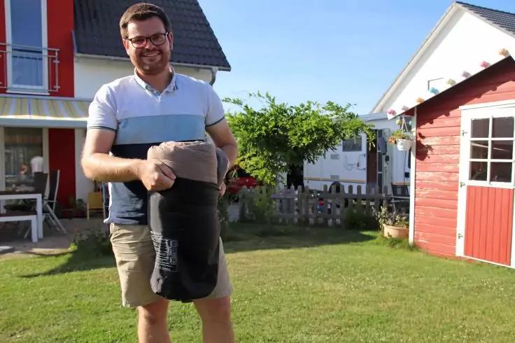 Mann steht auf Rasen vor zwei H&auml;usern und h&auml;lt eine in einer schwarzen Trage befindliche Person