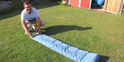 Person mit Brille kniet auf dem Rasen und rollt einen hellblauen Schlafsack vor einem roten Gartenschuppen aus