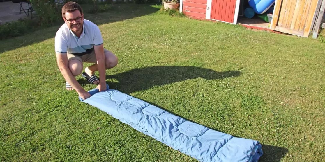 Person mit Brille kniet auf dem Rasen und rollt einen hellblauen Schlafsack vor einem roten Gartenschuppen aus