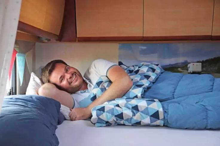 Ein Mann liegt seitlich auf einer Matratze, st&uuml;tzt den Kopf auf die Hand, ist zur H&auml;lfte unter einer blau gemusterten Decke, auf einem hellblauen Kissen, im Innenraum mit h&ouml;lzernen Oberschr&auml;nken und einem Fenster mit Vorhang; an der Wand ein Poster m