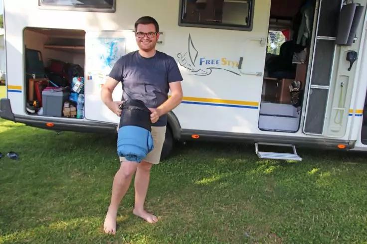 Eine Person steht barfu&szlig; auf Gras vor einem Wohnmobil und h&auml;lt ein Kind kopf&uuml;ber an den Beinen; ge&ouml;ffnete Wohnmobilt&uuml;r und offenes Staufach sichtbar