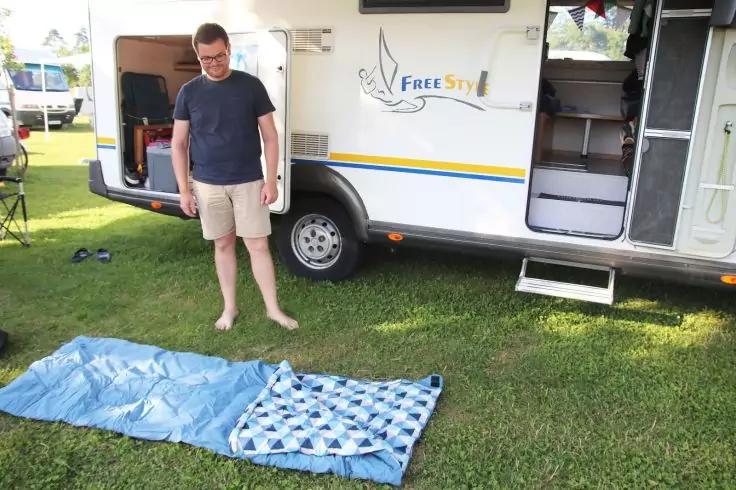 Ein Mann steht barfu&szlig; auf Gras neben einem ge&ouml;ffneten Wohnwagen; zwei Decken liegen auf dem Gras