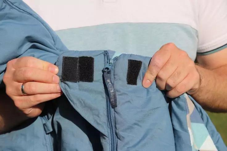 Zwei H&auml;nde halten eine hellblaue Jacke mit offenem Rei&szlig;verschluss und zwei schwarzen Klettverschl&uuml;ssen; im Hintergrund wei&szlig;es T‑Shirt mit hellblauem Streifen; Ring am linken Ringfinger