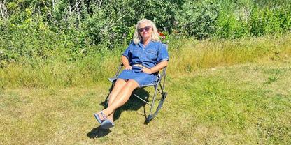 Person mit blondem Haar und Sonnenbrille sitzt in einem Klappstuhl auf einer Wiese vor Str&auml;uchern, tr&auml;gt blaues Kleid