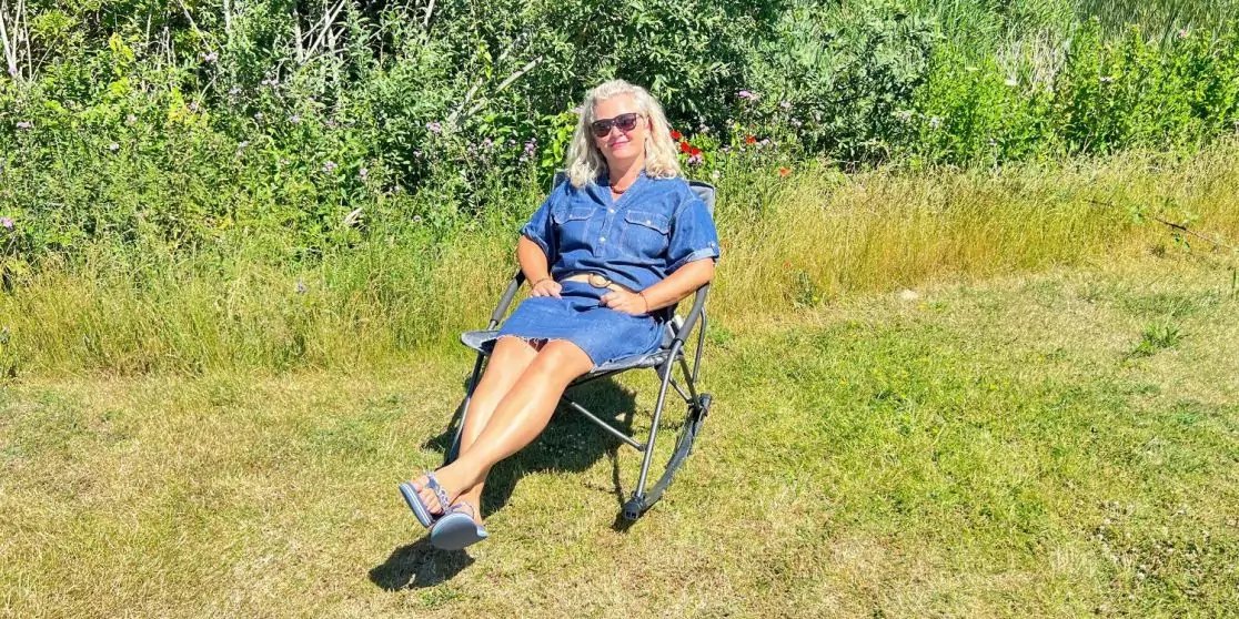 Person mit blondem Haar und Sonnenbrille sitzt in einem Klappstuhl auf einer Wiese vor Str&auml;uchern, tr&auml;gt blaues Kleid