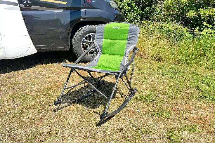 Gr&uuml;n-grauer klappbarer Schaukel-Campingstuhl auf Gras neben dem Rad und der Seitenwand eines wei&szlig;en Wohnmobils, dahinter B&uuml;sche