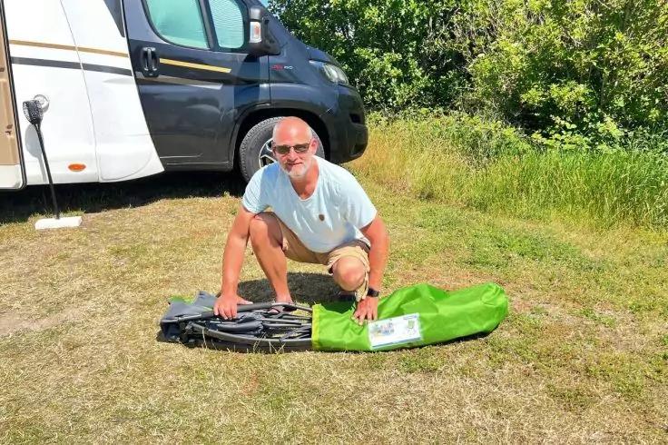 Mann kniet auf Gras, H&auml;nde auf einer gr&uuml;nen und einer schwarzen Tasche, geparktes Wohnmobil daneben, B&auml;ume im Hintergrund