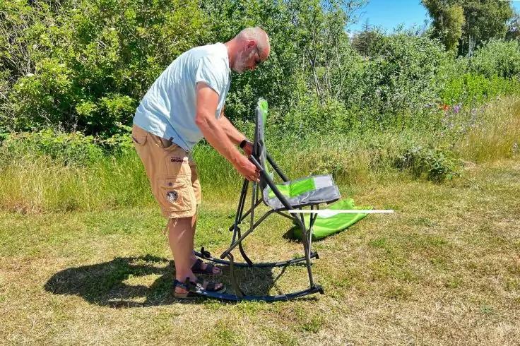 Erwachsener Mann mit Glatze und Bart in T‑Shirt und Shorts beugt sich &uuml;ber einen gr&uuml;nen Klappstuhl und h&auml;lt dessen Gestell; Grasfl&auml;che und B&uuml;sche im Hintergrund