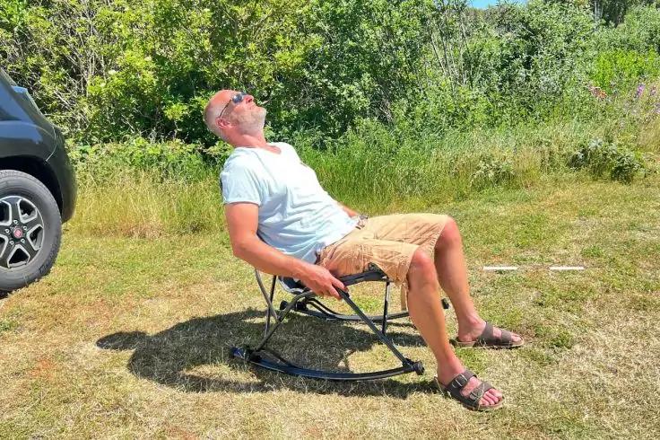 Ein Mann sitzt in einem klappbaren Schaukelstuhl auf trockenem Gras neben einem geparkten Auto, tr&auml;gt T-Shirt, Shorts, Sandalen und Sonnenbrille, lehnt sich mit zur&uuml;ckgelegtem Kopf zur&uuml;ck