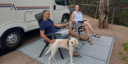 Zwei Personen sitzen auf Klappst&uuml;hlen auf einer gemusterten Matte neben einem wei&szlig;en Wohnmobil; ein Hund steht neben ihnen, zwei Becher auf einem kleinen Tisch