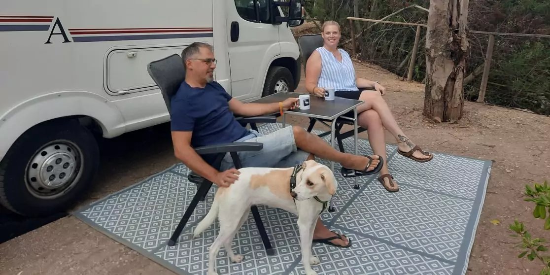 Zwei Personen sitzen auf Klappst&uuml;hlen auf einer gemusterten Matte neben einem wei&szlig;en Wohnmobil; ein Hund steht neben ihnen, zwei Becher auf einem kleinen Tisch