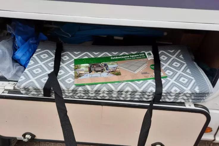 Offenes Fach mit zusammengerollter grau-weißer Matte mit Rautenmuster, zwei schwarze Gurte, grünes Produktetikett, blaue Plastiktüte links