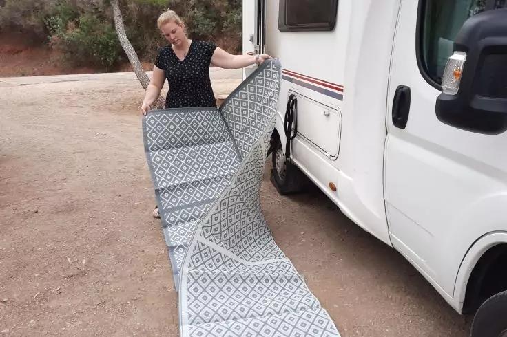 Person steht neben einem Wohnmobil und rollt einen gemusterten Teppich auf dem Boden aus