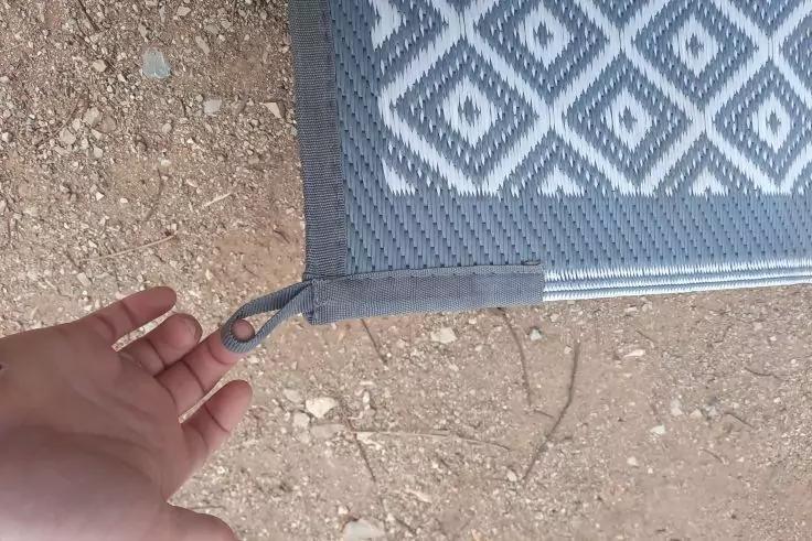 Hand hält Schlaufe einer grauen Matte mit weißem Diamantmuster auf kiesigem Boden