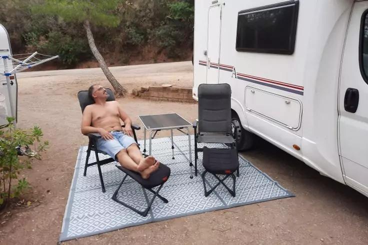 Person ohne Oberteil in Shorts liegt auf einem zusammenklappbaren Liegestuhl auf einer gemusterten Außenmatte neben einem weißen Wohnmobil; zwei Klappstühle und ein kleiner Tisch; sandiger Boden, Bäume im Hintergrund