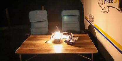 Holztisch mit einer brennenden Laterne in der Mitte, zwei gepolsterte Campingst&uuml;hle dahinter, auf dem Tisch eine T&uuml;te Chips und eine Zeitschrift; rechts eine Wand mit blauen und gelben Streifen