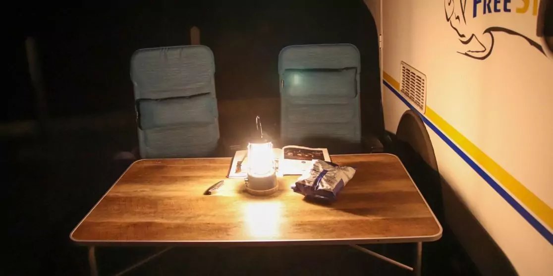 Holztisch mit einer brennenden Laterne in der Mitte, zwei gepolsterte Campingst&uuml;hle dahinter, auf dem Tisch eine T&uuml;te Chips und eine Zeitschrift; rechts eine Wand mit blauen und gelben Streifen