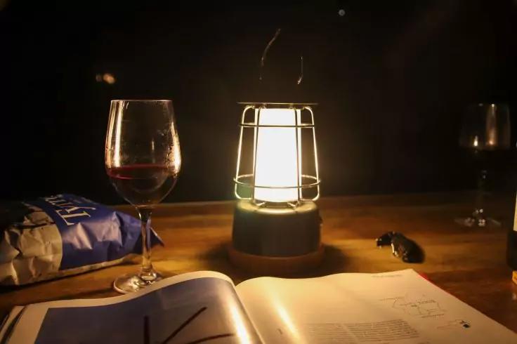 Auf einem Holztisch eine brennende Laterne in der Mitte, links ein halb gefülltes Rotweinglas und eine geöffnete Chipstüte, rechts ein weiteres Rotweinglas und ein kleiner Gegenstand; vorne ein aufgeklapptes Buch mit sichtbaren Seiten, dunkler Hinter