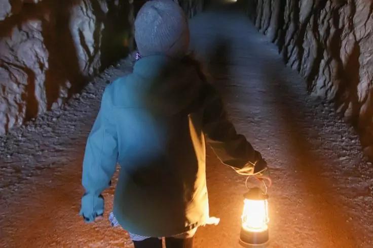 Person von hinten in Kapuzenjacke hält eine Laterne in einem engen Tunnel mit rauen Wänden und unbefestigtem Boden