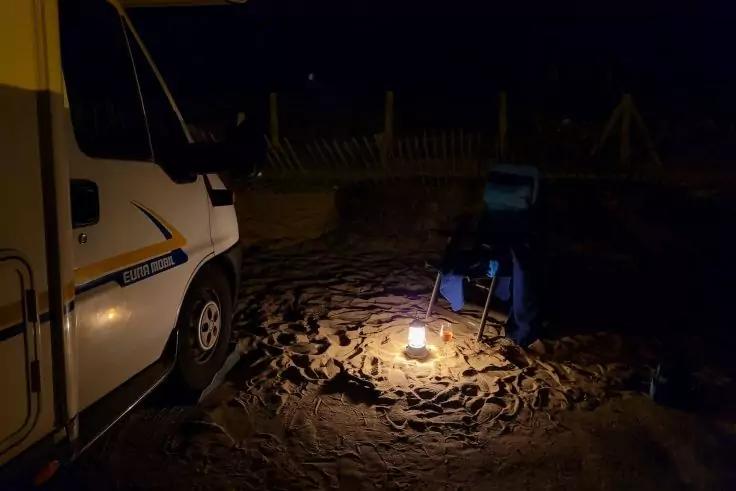 Teil eines Wohnmobils links, sandiger Boden mit Spuren, kleine Laterne auf dem Sand, blauer Klappstuhl und kleiner Beistelltisch rechts, dunkler Hintergrund mit Zaun