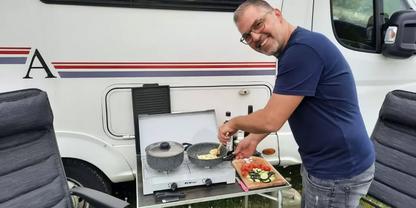 Mann steht neben Wohnmobil und kocht auf einem Zweiflamm-Gaskocher auf einem Klapptisch; Schneidebrett mit geschnittenem Gem&uuml;se und zwei Campingst&uuml;hle sichtbar