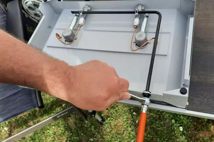Eine Hand h&auml;lt einen verstellbaren Schraubenschl&uuml;ssel an der Verbindung zwischen einem orangefarbenen Gasschlauch und einem schwarzen Metallrohr eines tragbaren grauen Gaskochergeh&auml;uses mit zwei Brennern, Gras im Hintergrund