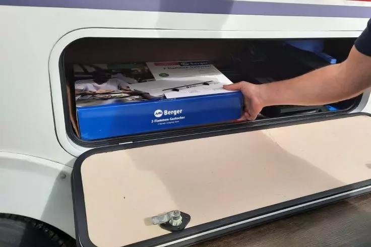 Arm schiebt eine blaue Box mit wei&szlig;em "Berger"-Logo in ein offenes seitliches Fach; ge&ouml;ffnete beige Klappe und Teil eines Rads sichtbar