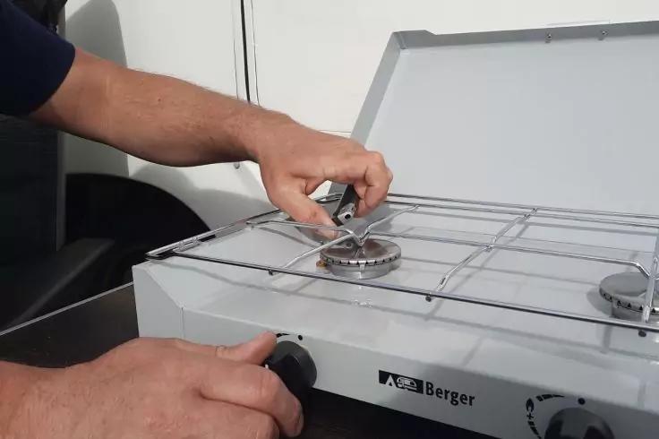 H&auml;nde platzieren ein Metallgitter &uuml;ber einem Brenner eines wei&szlig;en mobilen Gaskochers; eine Hand greift einen vorderen Drehknopf; Geh&auml;use mit Schriftzug "Berger" sichtbar