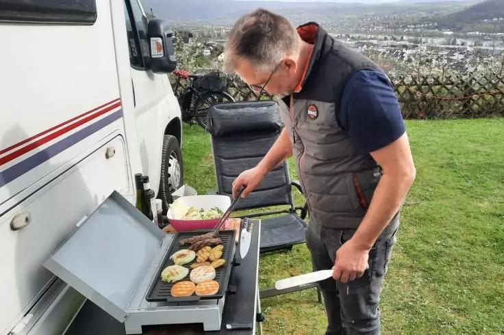 Mann steht neben geparktem Wohnmobil und wendet Br&ouml;tchen und Gem&uuml;se auf einer aufklappbaren Grillplatte; Sch&uuml;ssel mit Salat, Klappstuhl, Rasen und Stadt im Hintergrund