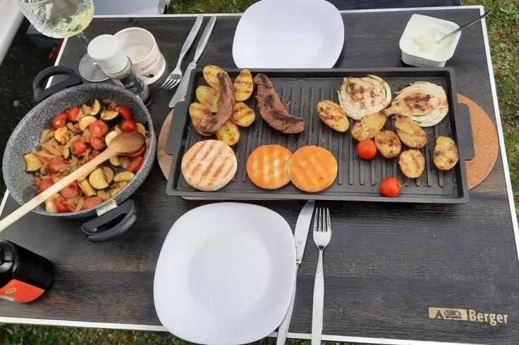 Grillplatte auf Tisch mit gebr&auml;unten Kartoffelh&auml;lften, drei runden orange-braunen Scheiben, l&auml;nglichem braunen St&uuml;ck und Kirschtomaten; Pfanne mit geschnittenem Gem&uuml;se, wei&szlig;e Teller, Besteck, Saucensch&auml;lchen und Glas auf Rasen