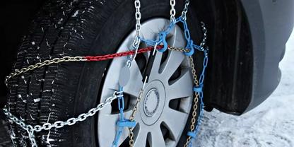 Nahaufnahme eines Autoreifens mit Schneeketten, blauen Spannern und einer roten Leine, Schnee auf dem Boden