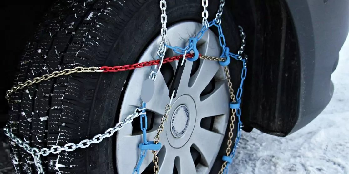 Nahaufnahme eines Autoreifens mit Schneeketten, blauen Spannern und einer roten Leine, Schnee auf dem Boden
