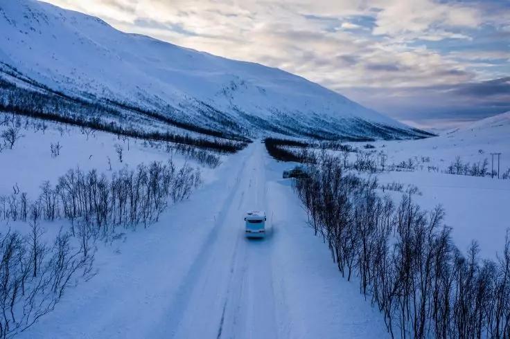 Ein Auto auf einer schneebedeckten Stra&szlig;e zwischen niedrigen kahlen B&uuml;schen, umgeben von verschneiten Bergen und einem bew&ouml;lkten Himmel