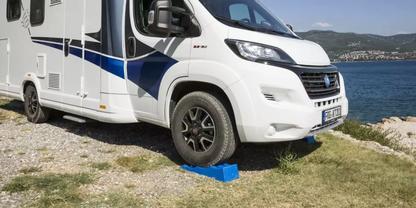 Wei&szlig;es Wohnmobil am Ufer auf Kies und Gras, blaues Ausgleichskeil unter dem vorderen Rad, im Hintergrund Wasser und entfernte K&uuml;ste mit Geb&auml;uden und H&uuml;geln