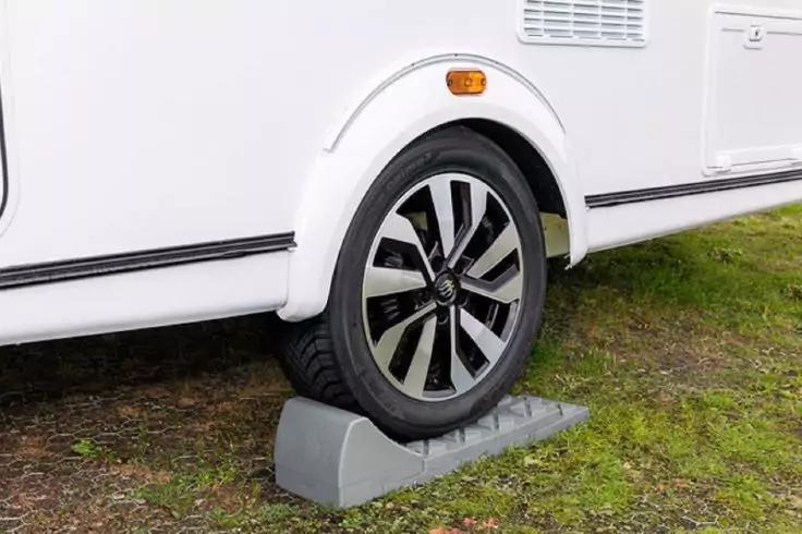 Rad eines wei&szlig;en Wohnwagens auf grauem Unterlegkeil, gr&uuml;ner Rasen, orangefarbener Seitenreflektor