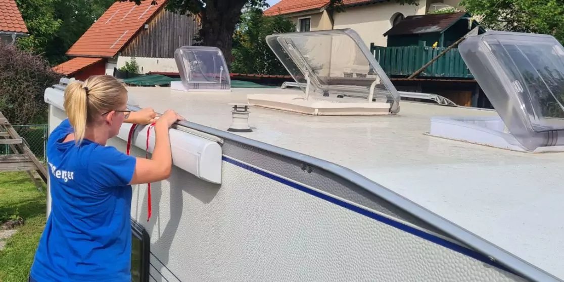 Person mit blondem Pferdeschwanz in blauem T‑Shirt steht seitlich an einem wei&szlig;en Wohnwagen, H&auml;nde an der Seitenwand; auf dem Dach zwei ge&ouml;ffnete Dachluken; im Hintergrund H&auml;user mit roten D&auml;chern und Gr&uuml;n