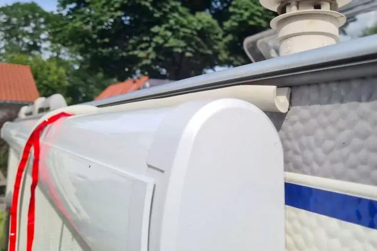 Weißes, abgerundetes Kunststoffgehäuse an Metallprofil, darunter strukturierte weiße Fläche mit blauem Streifen; rotes Band links; Bäume und Hausdach im Hintergrund