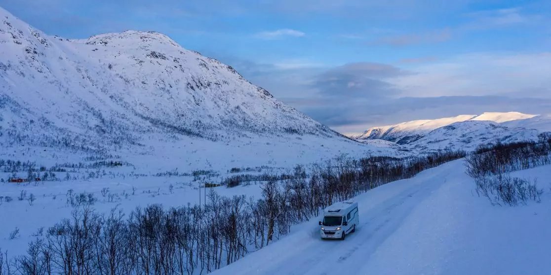 Wei&szlig;er Transporter auf schneebedeckter Stra&szlig;e in verschneitem Tal, kahle B&uuml;sche, schneebedeckte Berge, blauer Himmel mit Wolken