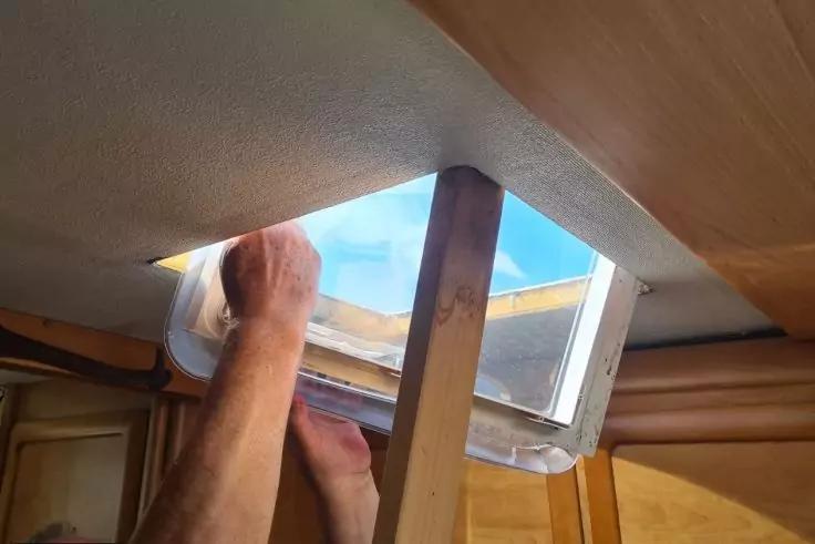 Zwei H&auml;nde am Rahmen eines ge&ouml;ffneten rechteckigen Deckenfensters; blauer Himmel durch die &Ouml;ffnung; Holzverkleidung und ein vertikaler Holzbalken sichtbar