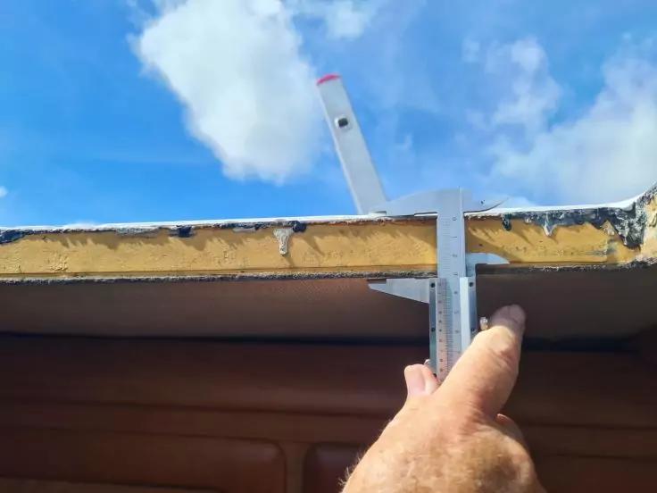 Hand h&auml;lt Metall-Schieblehre an die Kante eines horizontalen Paneels mit gelber Schicht; darunter braune Innenfl&auml;che; blauer Himmel mit Wolken im Hintergrund