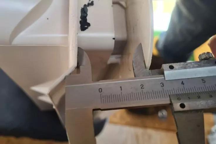 Messschieber mit ge&ouml;ffneten Backen an einem wei&szlig;en Kunststoffteil, sichtbare Skala mit Zahlen, kleine schwarze R&uuml;ckst&auml;nde auf dem Kunststoff, verschwommener Hintergrund mit Person und Holzboden