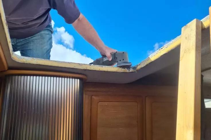 Person in Jeans steht an einer ge&ouml;ffneten Dachluke und h&auml;lt ein Schleifger&auml;t &uuml;ber einem Holzrahmen; blauer Himmel mit Wolken