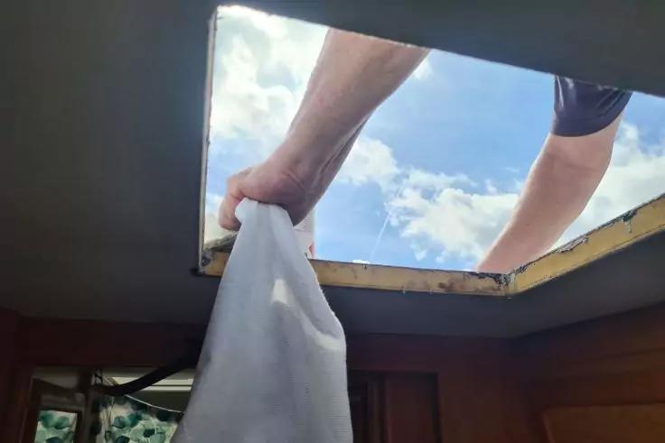 Zwei ausgestreckte Arme durch eine rechteckige Dach&ouml;ffnung, die ein wei&szlig;es Tuch halten; blauer Himmel mit Wolken durch die &Ouml;ffnung; Deckenrand und Holzverkleidung im Innenraum sichtbar