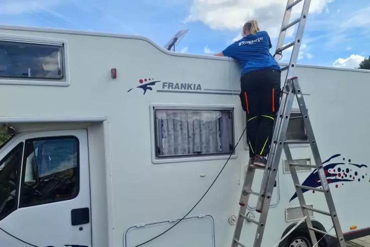 Person auf Leiter an der Seite eines wei&szlig;en Wohnmobils mit Schriftzug FRANKIA, Fenster mit Gardine, Kabel h&auml;ngt, blauer Himmel mit Wolken
