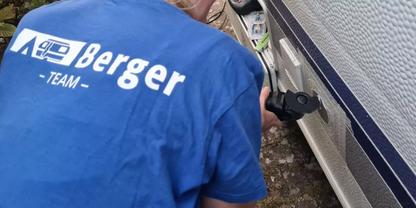 Person in blauem T-Shirt mit wei&szlig;em Aufdruck "Berger - TEAM", geb&uuml;ckt neben einem wei&szlig;en Wohnwagen mit blauer Zierlinie, h&auml;lt ein schwarzes Bauteil an der Au&szlig;enwand