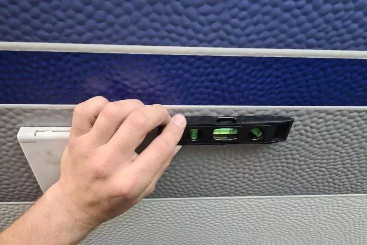 Linke Hand h&auml;lt eine schwarze Wasserwaage an eine zweifarbige Wand (blau oben, hellgrau unten) mit d&uuml;nnem wei&szlig;en Streifen und sichtbarem wei&szlig;en rechteckigem Bauteil